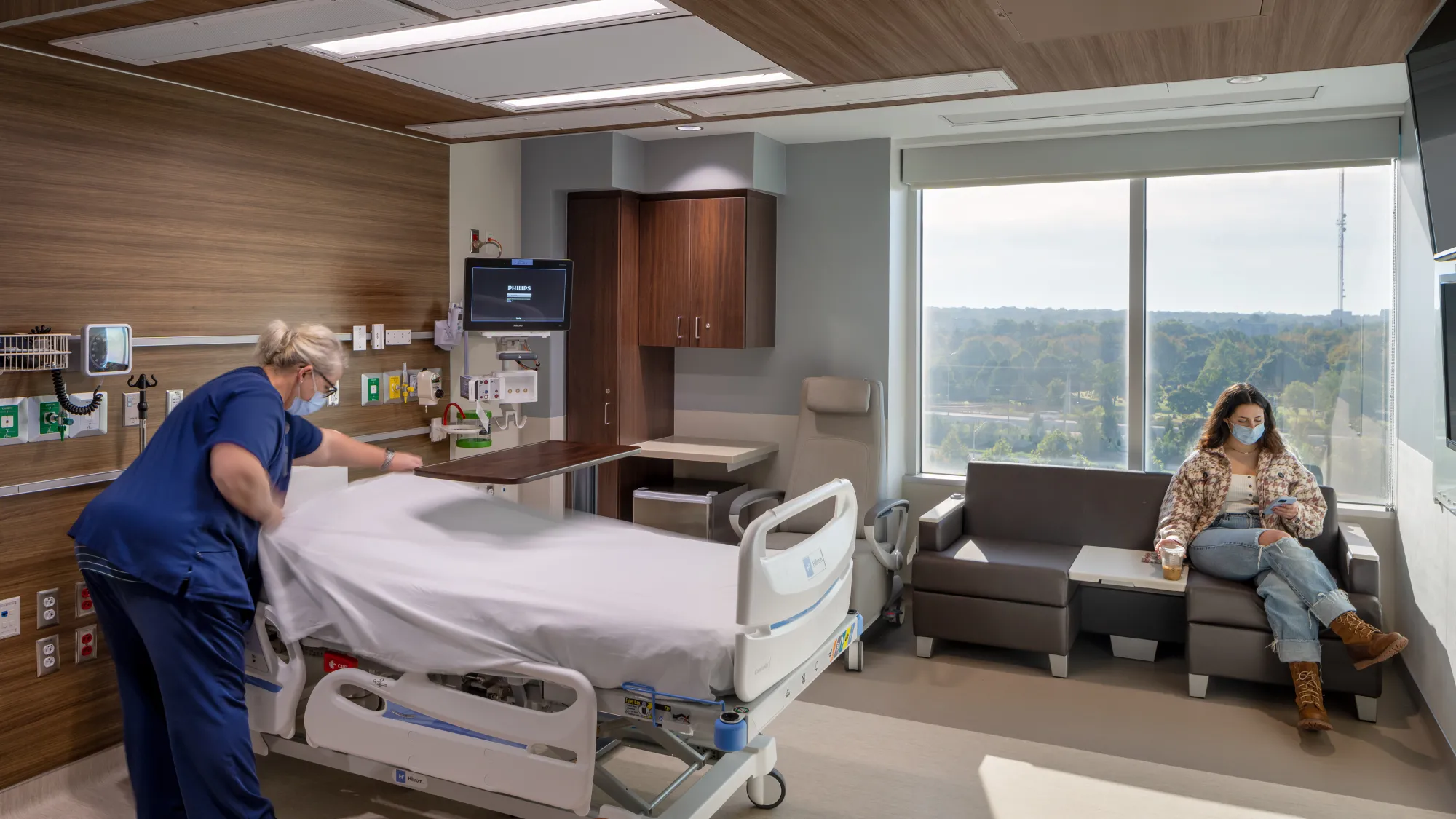 Interior day of a hospital room with a nurse making the bed and a woman sitting by the window on her phone