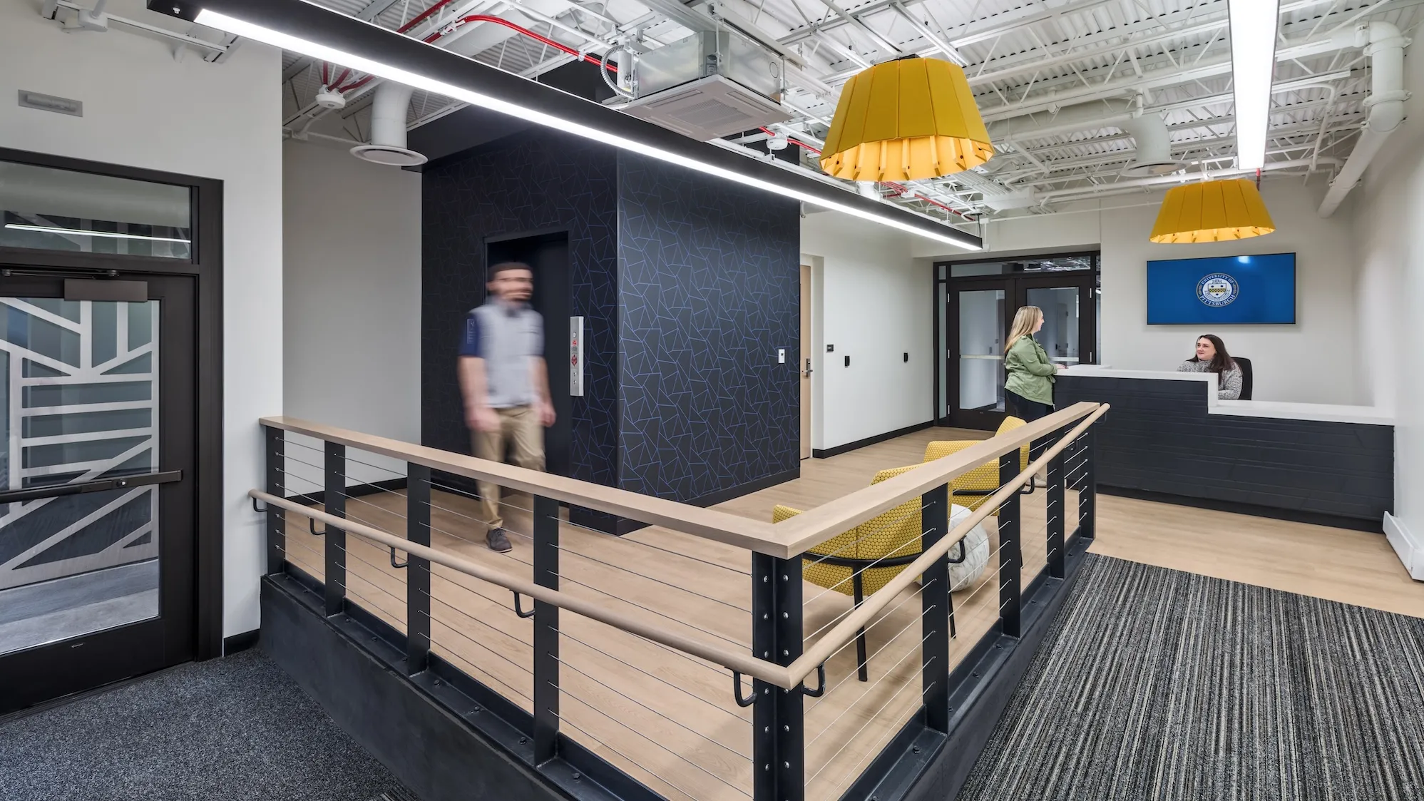 Interior of Pittsburgh University IT department entrance space, featuring a wooden ramp, front desk and yellow accented furniture and lights.