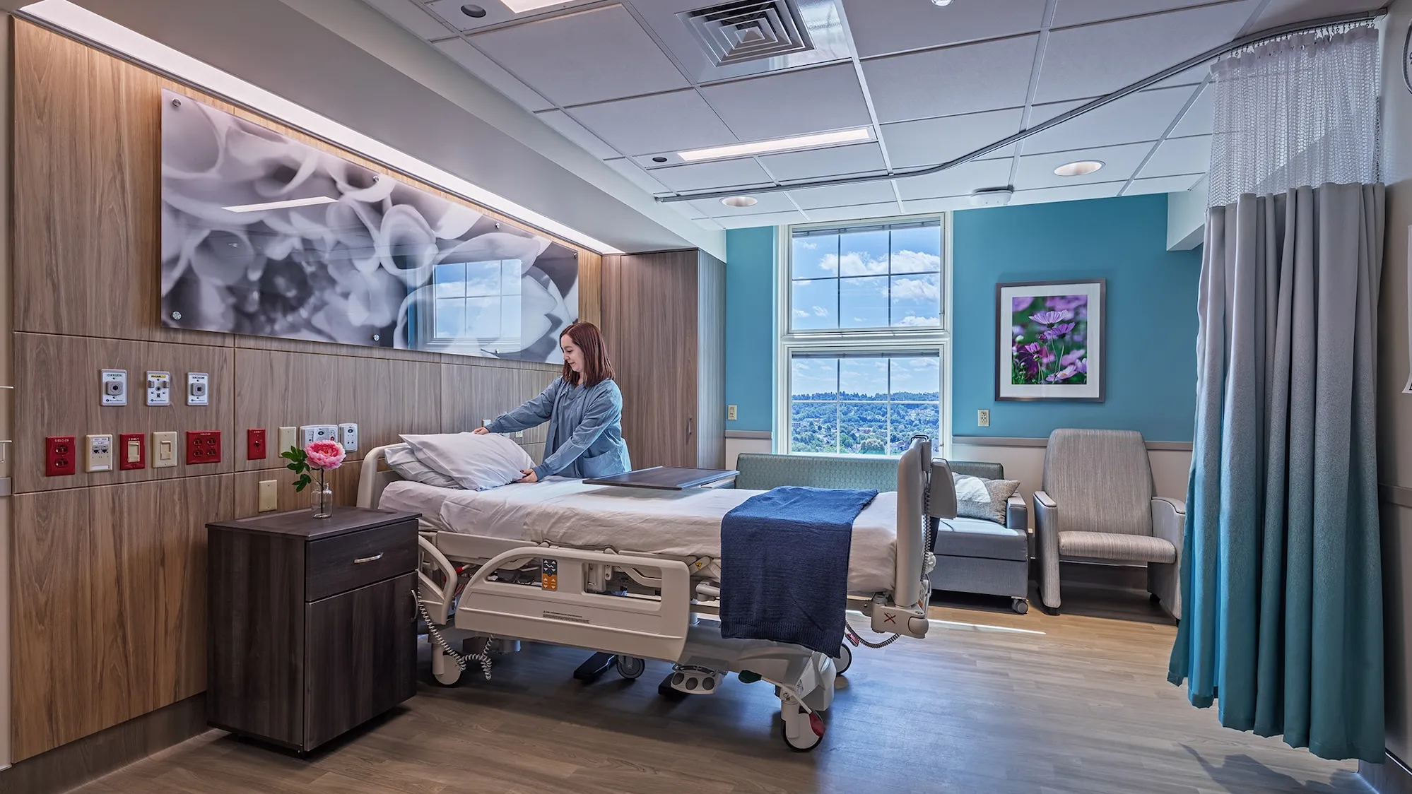 A nurse adjusting a bed pillow in a private hospital room, blue walls, floral art and window