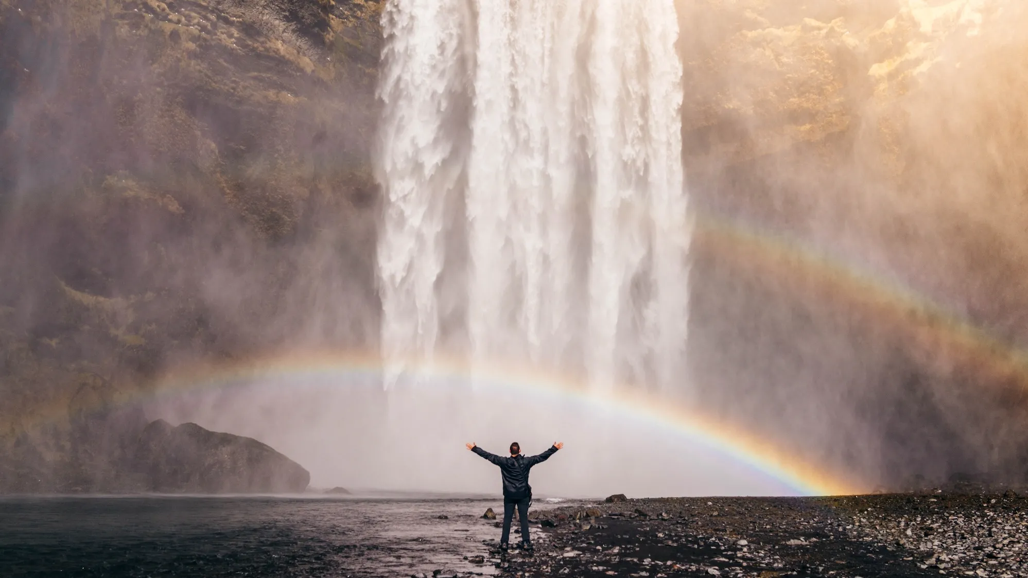 Person standing in front of a waterfall and two rainbows with their hands out and in the air.