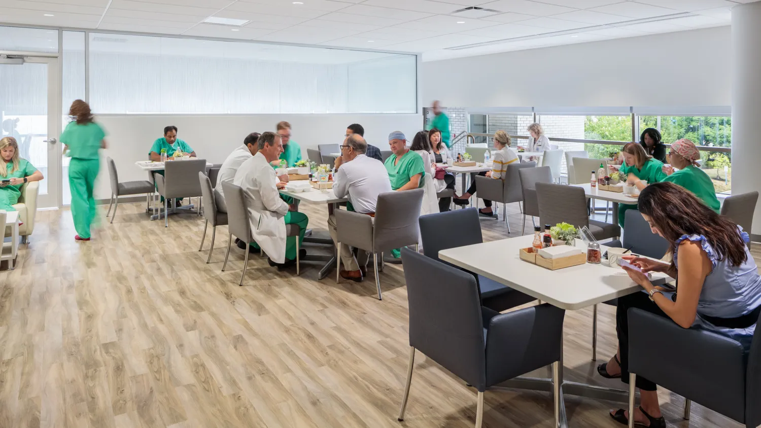 Cafeteria with many people in medical uniforms eating lunch, talking, and working at tables.