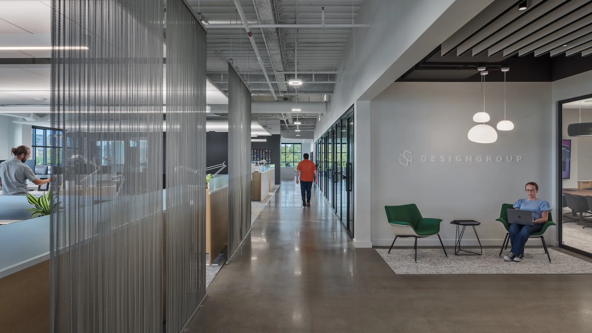 Office lobby with two green chairs, one person working on a laptop and the other walking down the lit hallway.