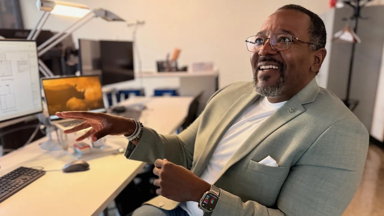 A man with glasses sitting at his desk gesturing with his hands while smiling.