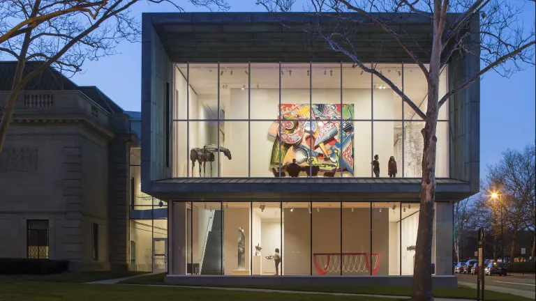 Exterior of the Columbus Museum of Art at night with the interior lights on, highlighting the floor to ceiling windows to peek into various colorful large art pieces