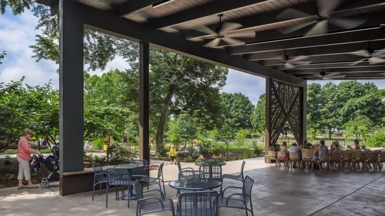 Outdoor large gazebo space with multiple ceiling fans and patio style tables surrounded by a variety of green plants and trees