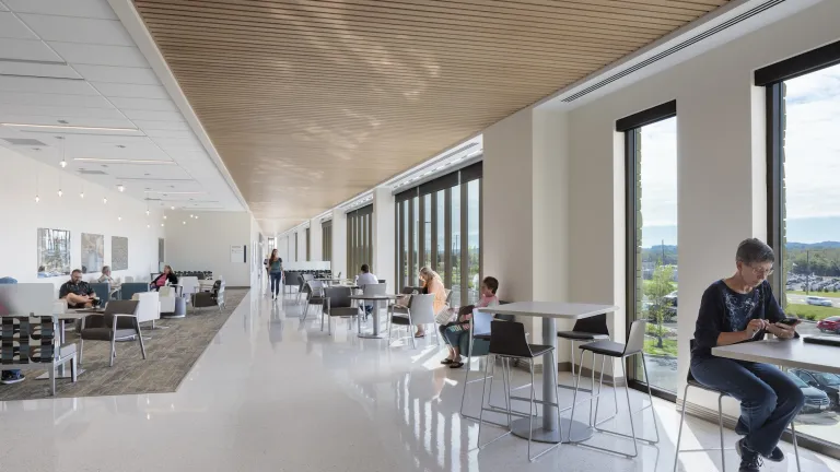 Upper floor open space with variety of low and tall tables and chairs on either side of a long concourse with floor to ceiling windows