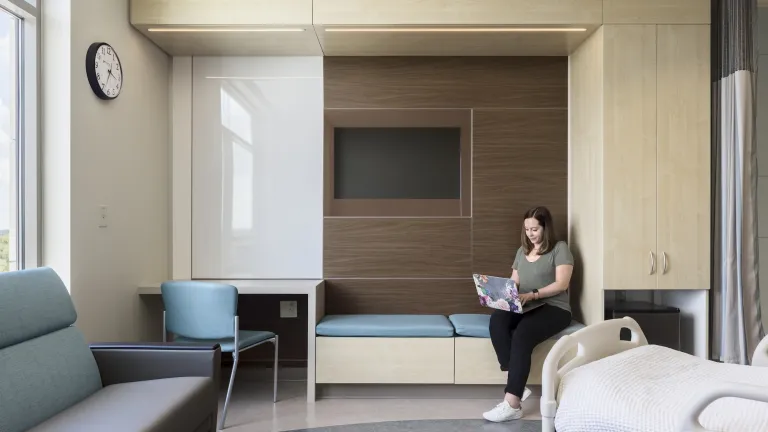 Interior day of a woman working on a laptop inside of a patient's room with accents of birch and light blue furnishings