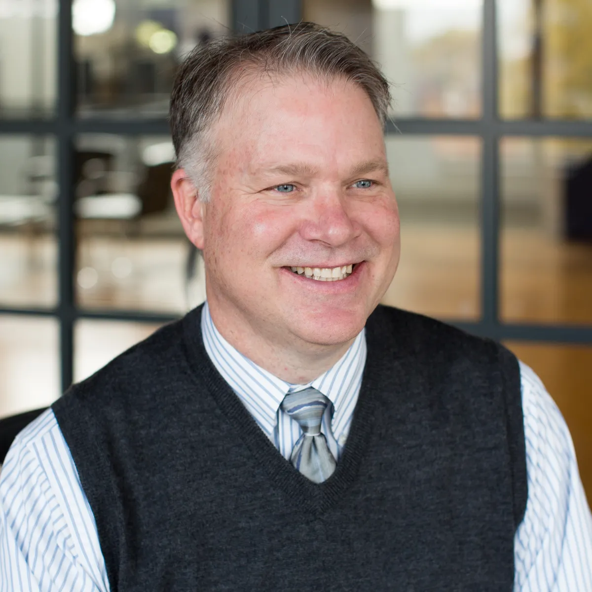 Mark Anderson smiling, looking off camera, wearing a dark gray sweater vest with light grey tie and button down shirt.