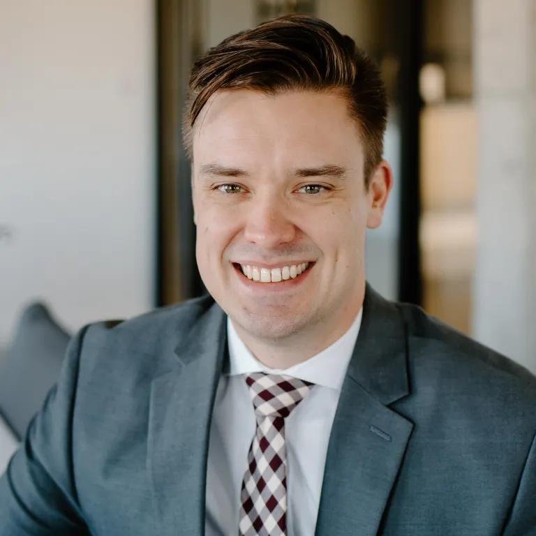 Joe Mayer smiling while wearing a grey blazer and gingham tie