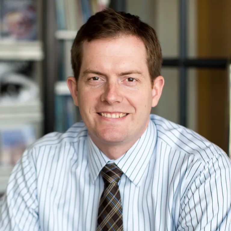 John Ryan smiling, wearing a white stripped button down and gingham tie