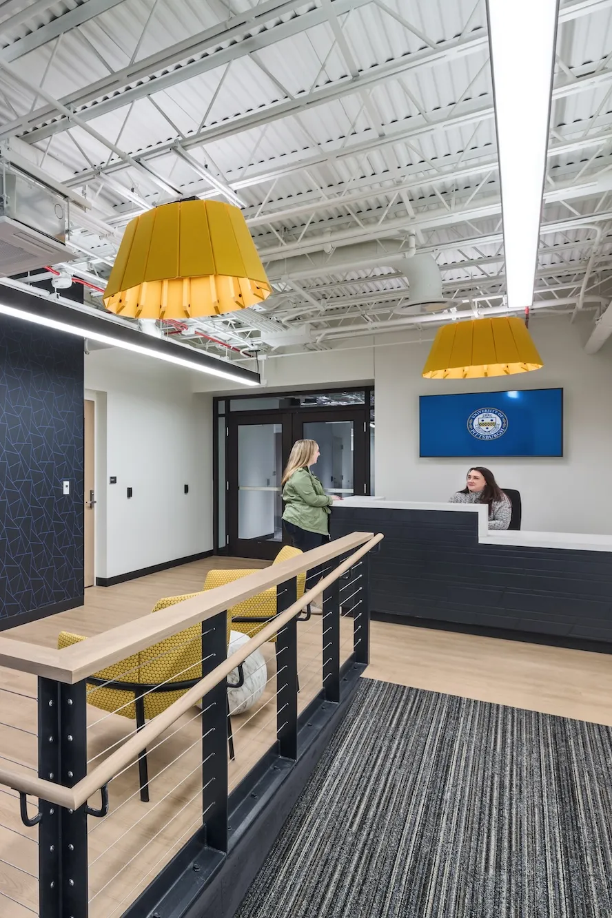 Interior of Pittsburgh University IT department entrance space, featuring a wooden ramp, front desk and yellow accented furniture and lights.