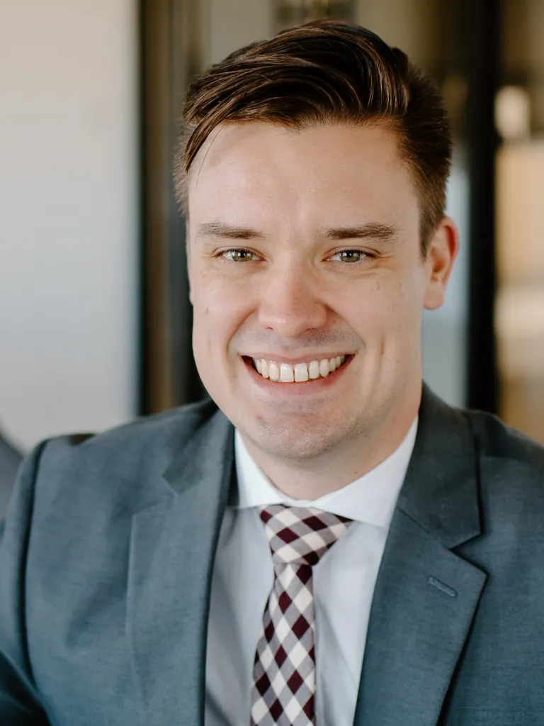 Joe Mayer smiling while wearing a grey blazer and gingham tie