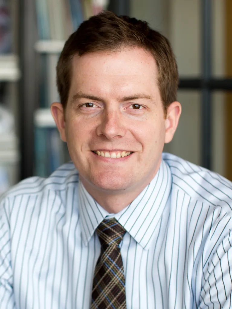 John Ryan smiling, wearing a white stripped button down and gingham tie