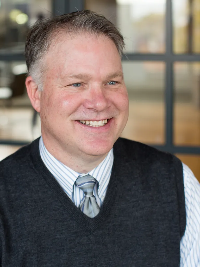 Mark Anderson smiling, looking off camera, wearing a dark gray sweater vest with light grey tie and button down shirt.