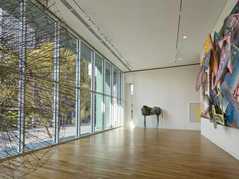 Interior of the Margaret M Walter wing, featuring floor to ceiling windows and three large art pieces, white walls and warm brown flooring