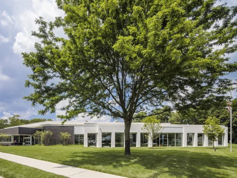 Exterior of Northern Lights Columbus Library during the day, a modern and gray one story building featuring a large green tree and a blue cloudy sky