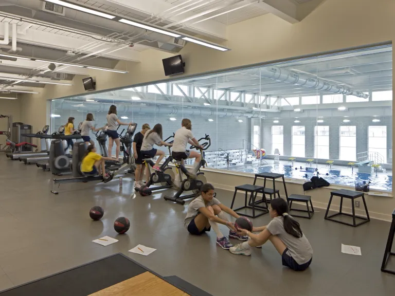 Second floor fitness space with a large window overlooking the swimming pool below, variety of girls working out on machines