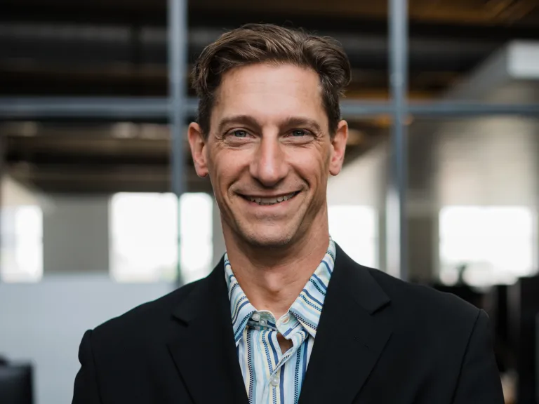 Dan Malec-Kosak smiling, wearing a black blazer and striped blue shirt