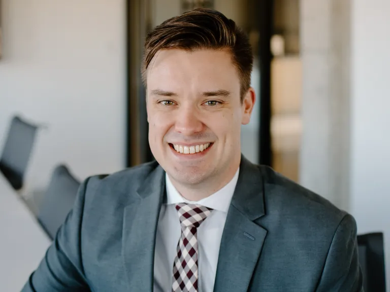 Joe Mayer smiling while wearing a grey blazer and gingham tie