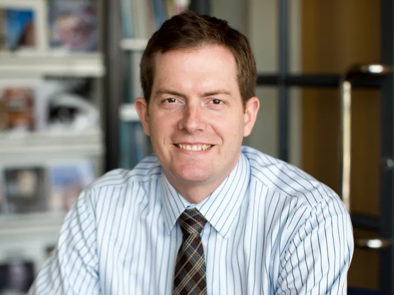 John Ryan smiling, wearing a white stripped button down and gingham tie