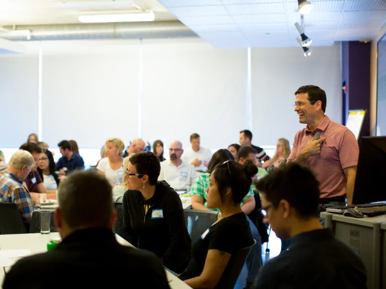 A conference room full of people, the speaker is smiling and pointing towards his chest