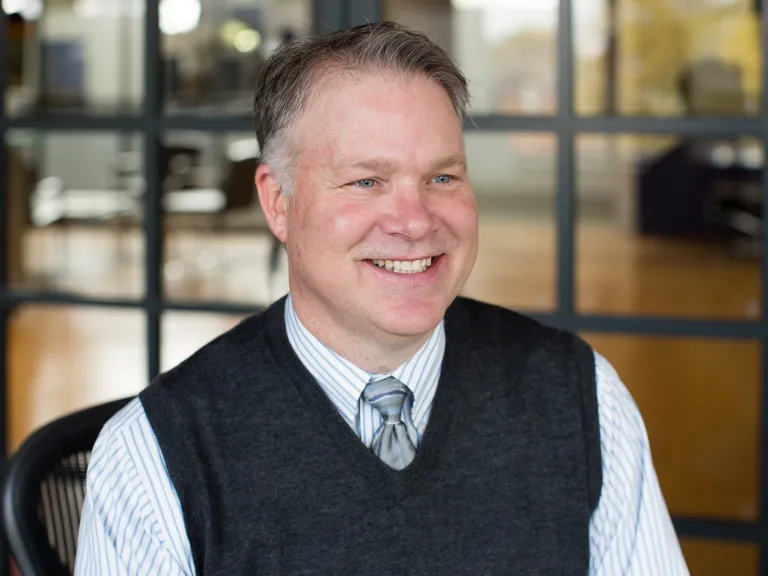 Mark Anderson smiling, looking off camera, wearing a dark gray sweater vest with light grey tie and button down shirt.