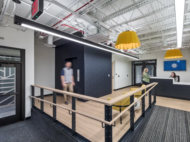 Interior of Pittsburgh University IT department entrance space, featuring a wooden ramp, front desk and yellow accented furniture and lights.