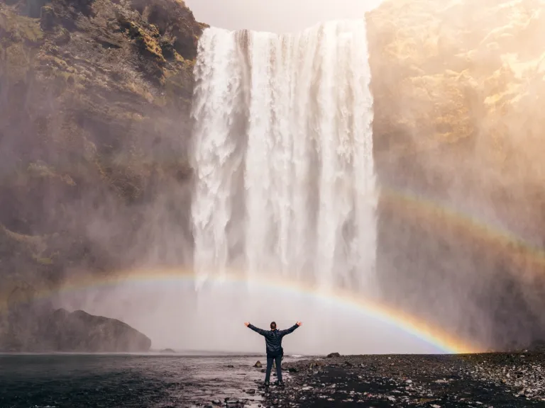 Person standing in front of a waterfall and two rainbows with their hands out and in the air.